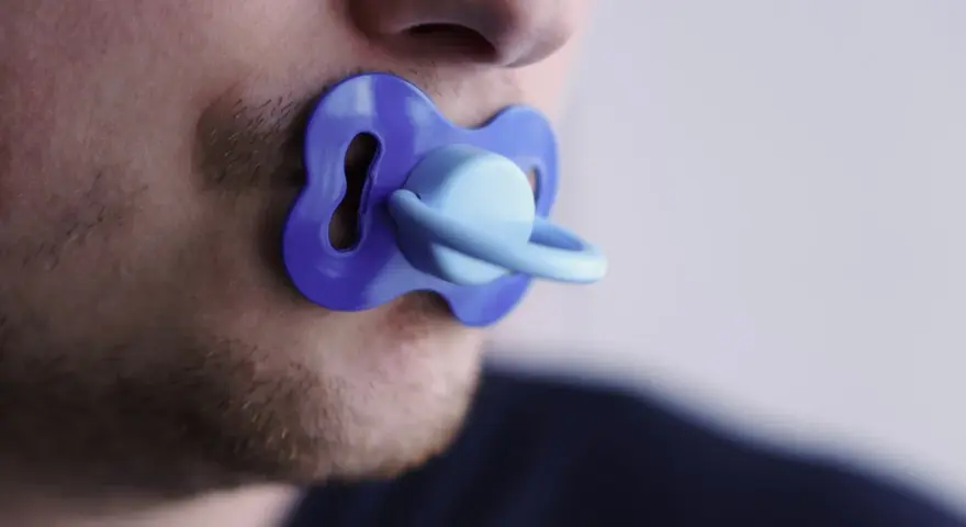 Un uomo con barba corta ha un ciuccio blu in bocca, un'immagine che evoca adulti che si comportano da bambini.