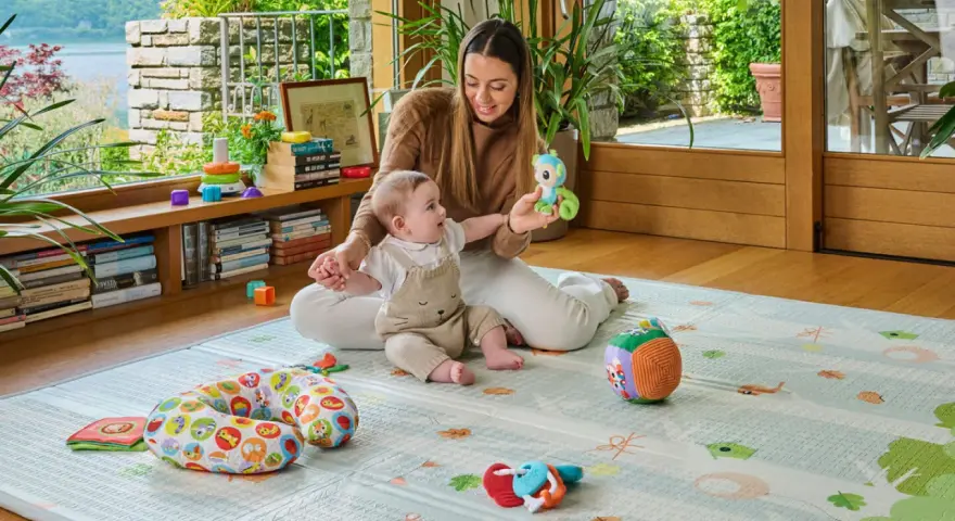 Mamma gioca con un neonato di 6 mesi che non sta seduto, su un tappeto gioco con tanti giocattoli colorati.