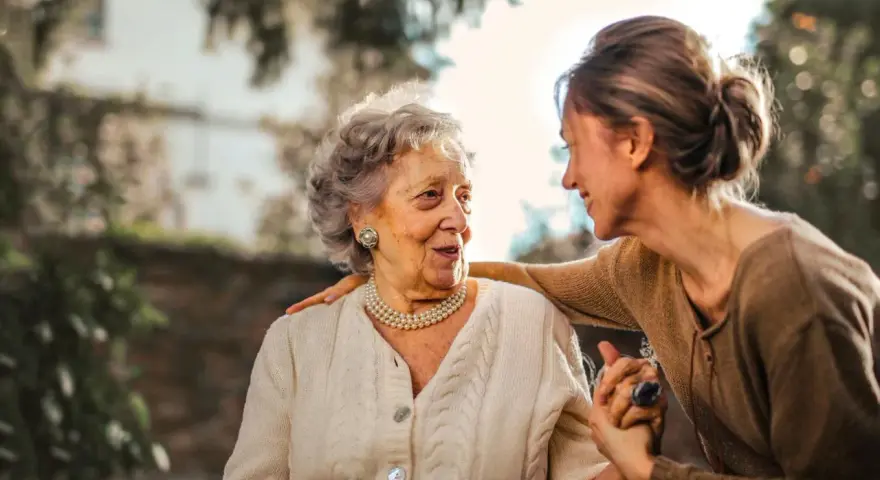 Una donna anziana e una giovane donna si guardano con affetto. La giovane donna ha una mano sulla spalla dell'anziana, un gesto che suggerisce cura e supporto, forse per superare le conseguenze di un cattivo rapporto con la madre.