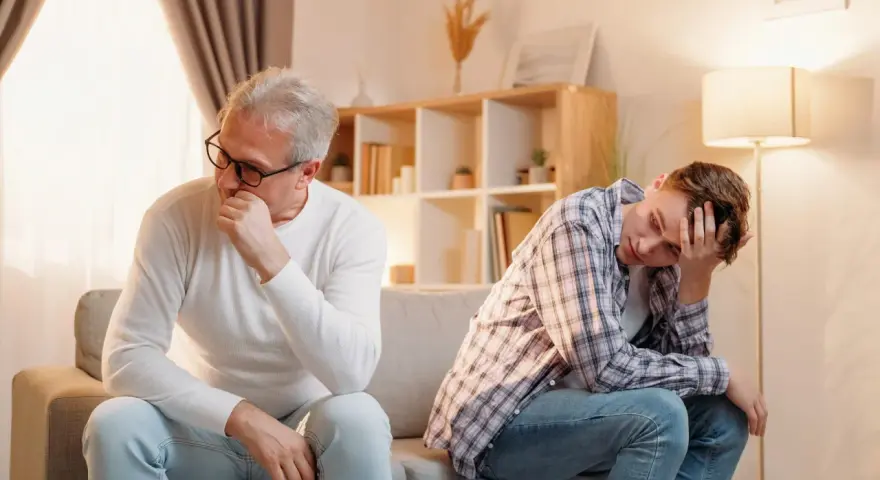 Padre e figlio seduti sul divano, distanti e pensierosi. Crescere in una famiglia dove si litiga sempre lascia segni profondi.