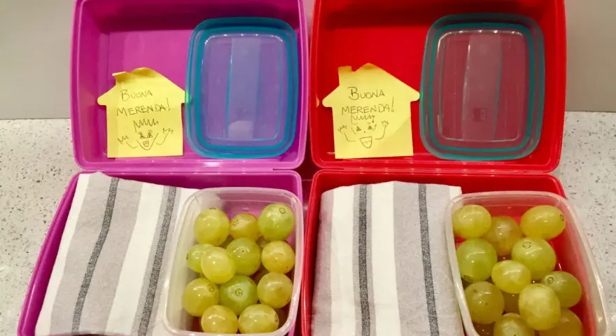Due lunchbox rosa e rosse pronte per le merende per bambini, con uva e un bigliettino "Buona Merenda!".