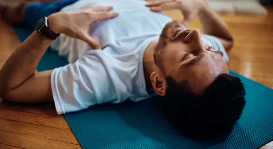 Uomo sdraiato su tappetino blu, mani sul petto, occhi chiusi. Pratica il training autogeno per gestire l'ansia.