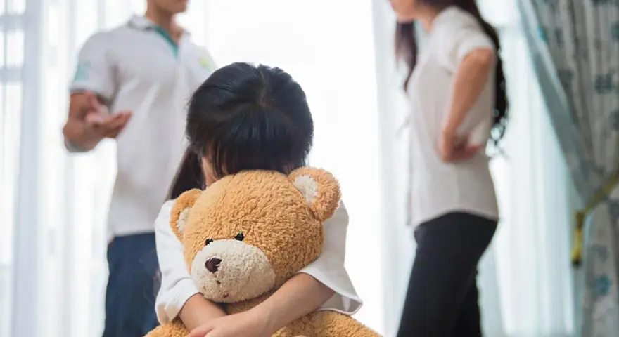 Bambina abbraccia orsacchiotto, mentre i genitori discutono. Avere coraggio di separarsi è difficile per tutti.