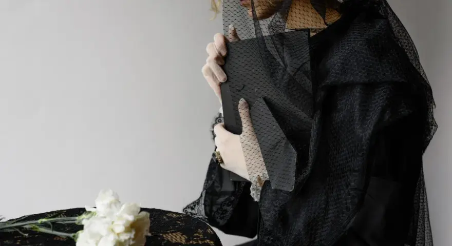 Donna velata di nero, con guanti bianchi, tiene un libro. Non accetto la morte di mio marito. Fiori bianchi su un tavolo coperto di pizzo nero.