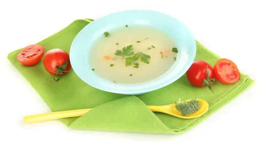 Brodo vegetale svezzamento: una ciotola azzurra con brodo, pomodorini e un cucchiaino giallo con broccoli.
