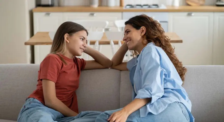 Madre e figlia si guardano sorridendo sul divano. La figlia, con un bottone mammario a 7 anni, ascolta attenta.