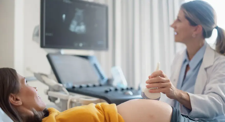 Ecografia di una donna incinta, con il medico che usa la sonda per sentire il battito del feto. L'emozione sul volto della futura mamma è palpabile.