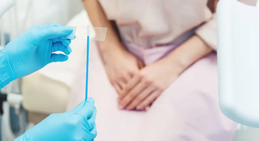 Mani con guanti blu tengono uno strumento per il pap test. La paziente, in attesa, è seduta.