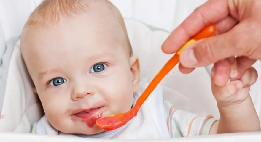 Bambino con occhi azzurri riceve la sua prima pappa svezzamento da un cucchiaino arancione.