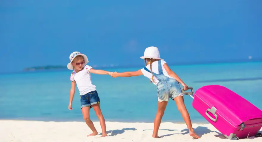 Due bambine con valigia rosa su spiaggia tropicale: idee per cosa fare a Ferragosto con i bambini.