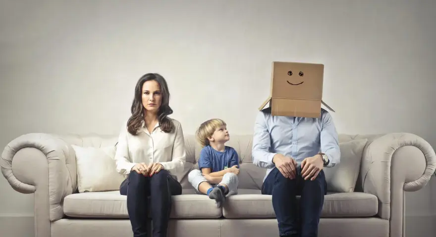 Famiglia seduta sul divano. La madre è triste, il bambino guarda il padre con la testa coperta da un cartone con un sorriso disegnato. Sembra che mio marito stia con me solo per i figli.