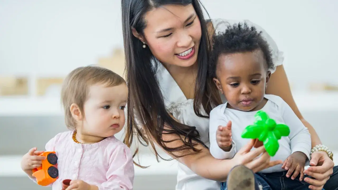 Bambini che giocano con giocattoli colorati, mentre una maestra sorride. Mi ricorda quando mio figlio ha iniziato a parlare a 4 anni.