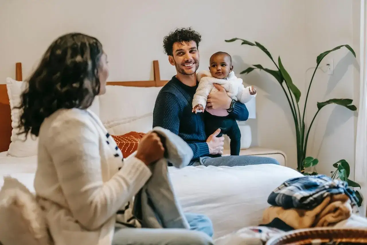 Un pap&agrave; sorridente tiene in braccio un neonato, mentre la mamma piega vestiti. L'arrivo di un fratellino a 3 anni porta gioia in famiglia.