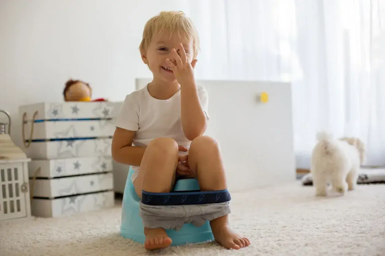 Bambino sorridente sul vasino, mentre il cane bianco si allontana. È una fase di apprendimento per i bambini, pipì in giro è un ricordo.