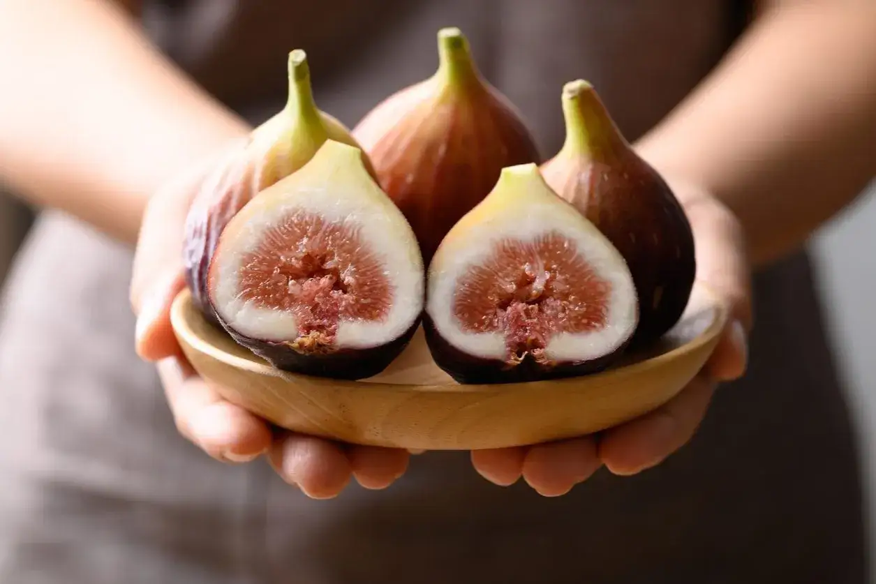 Mani che sorreggono un piattino di legno con fichi maturi, alcuni interi e uno tagliato a metà, mostrando la polpa succosa. Un'immagine che evoca la dolcezza e la generosità dei fichi in gravidanza.