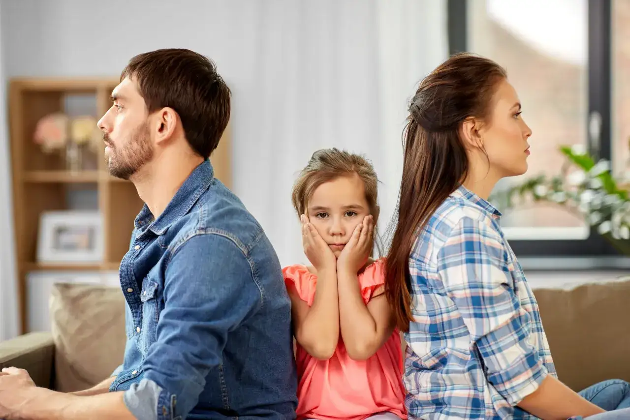Bambini con genitori separati, il cellulare è l'unico legame. La bambina è al centro, tra i genitori che si voltano le spalle, con le mani sul viso.