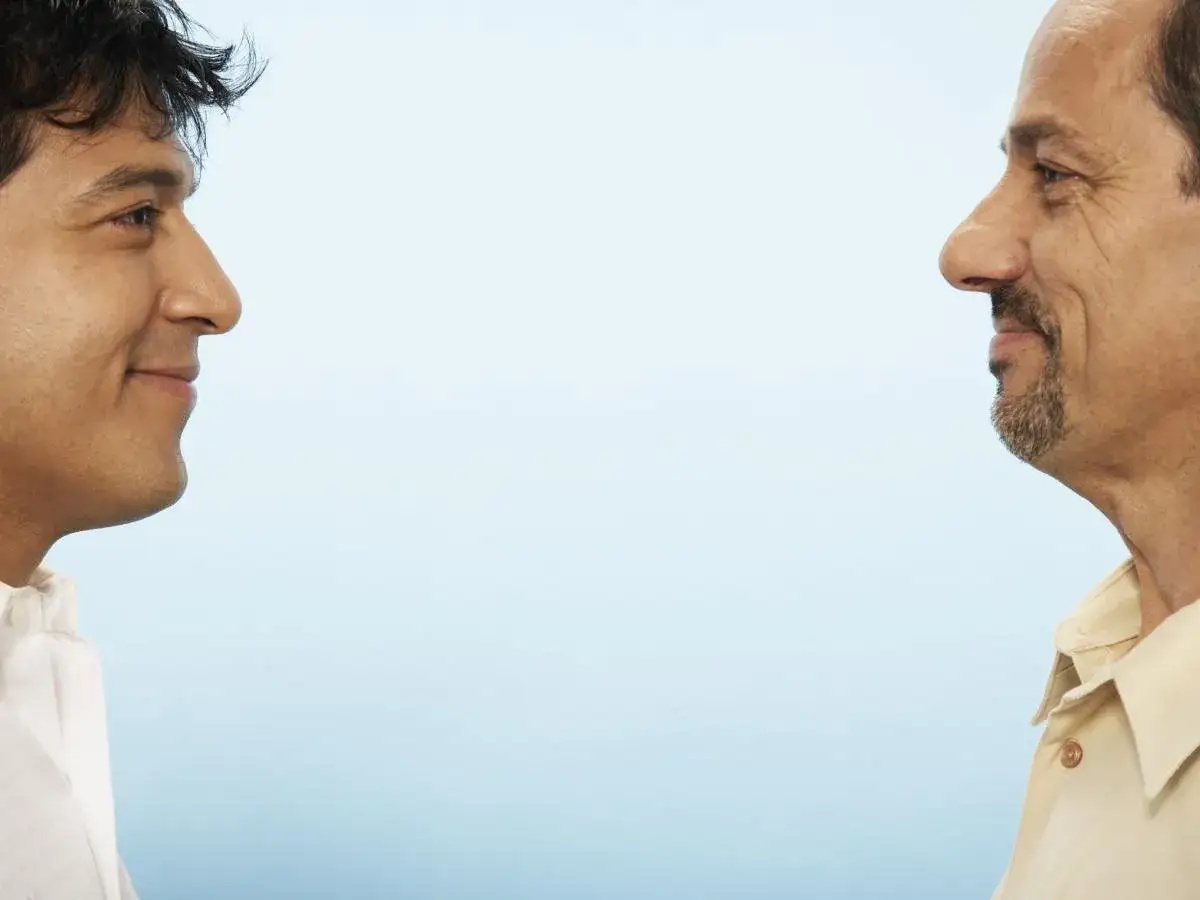 Due uomini si guardano, un sorriso timido sul volto del più giovane, mentre l'altro, con una leggera barba, sembra pensieroso. Un momento di attesa, forse un po' di ansia, prima di un confronto.