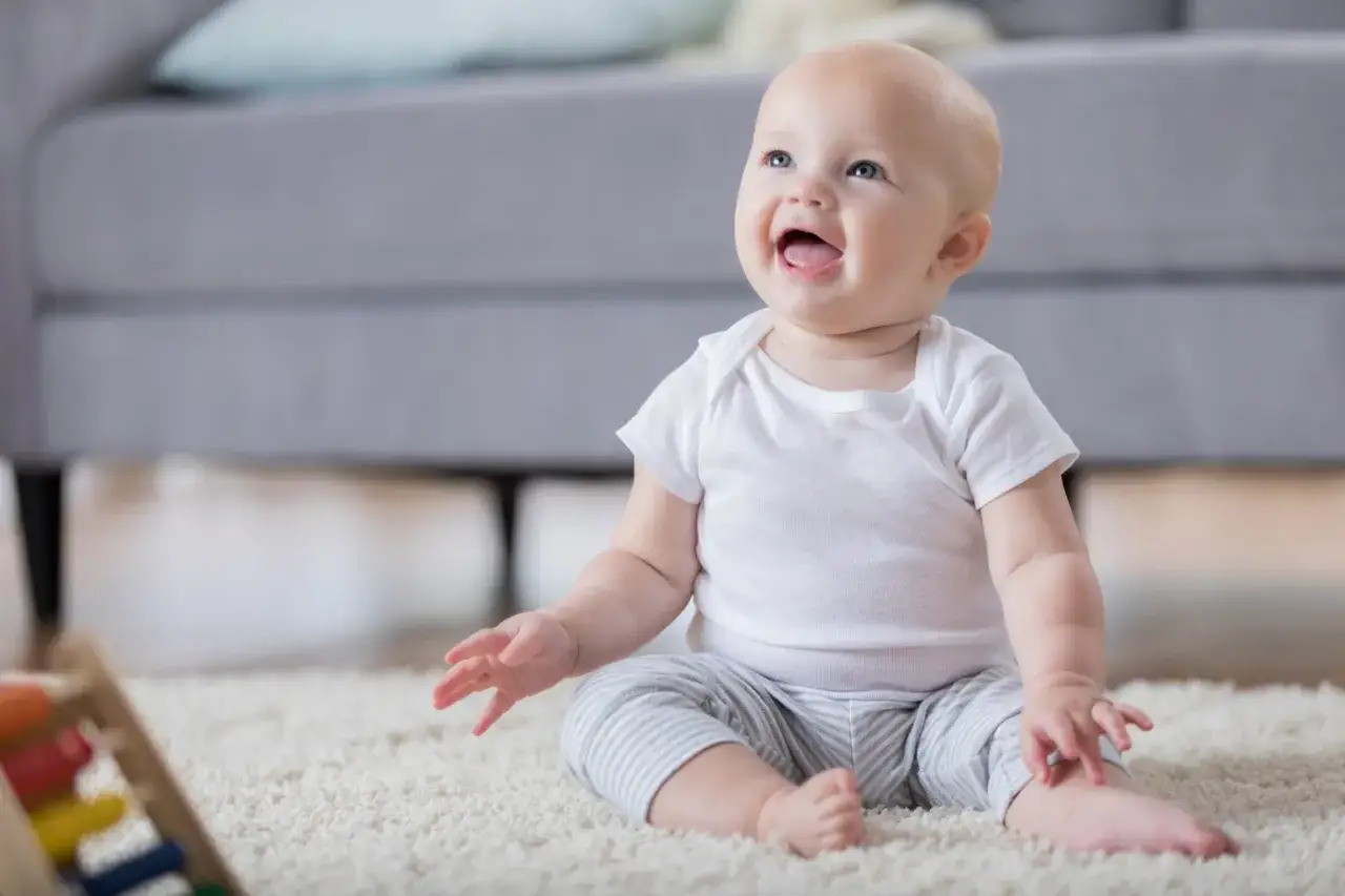 Bambino di 6 mesi, con un sorriso radioso, seduto sul tappeto. Non sta ancora seduto da solo, ma esplora il mondo con gli occhi.