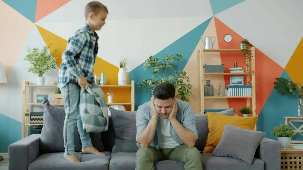 Un padre esausto si copre le orecchie mentre il figlio salta sul divano. Non sopporto pi&ugrave; i miei figli!
