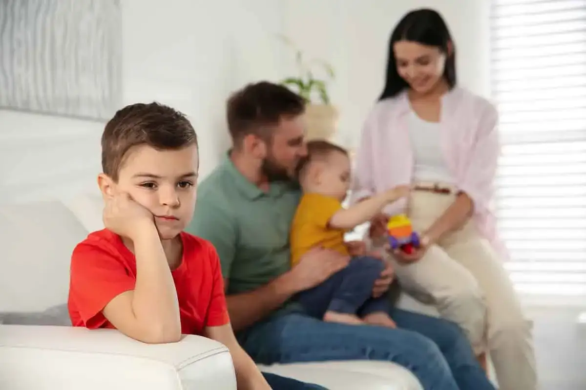 Un bambino con un'espressione malinconica sul divano, mentre i genitori coccolano il fratellino. Si percepisce la gelosia tra fratelli.