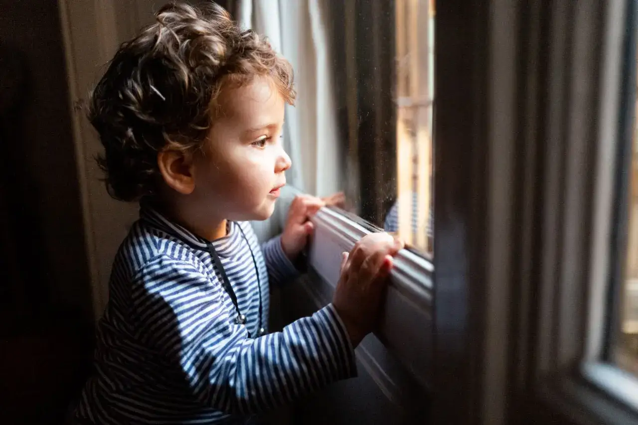 Bambino di 5 anni con riccioli guarda fuori dalla finestra, forse riflettendo sulle paure dei bambini.