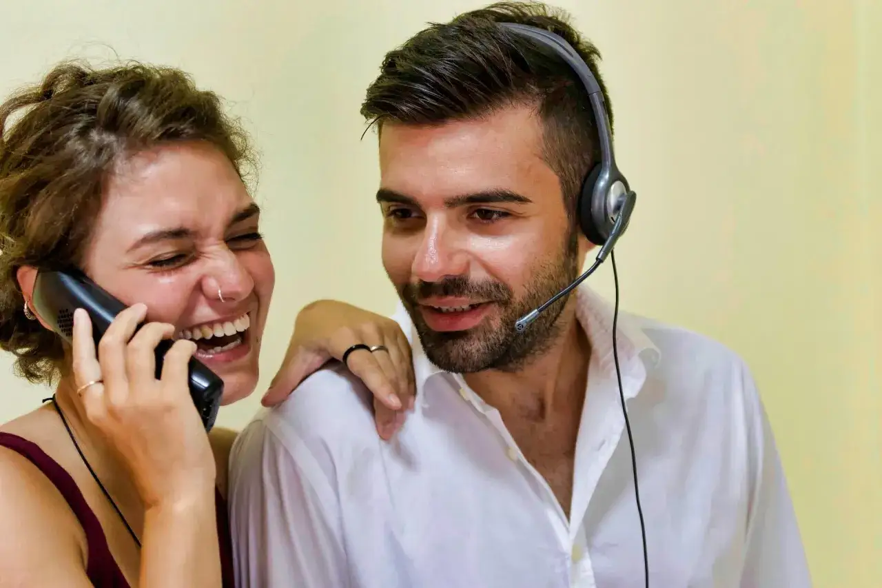Una donna ride al telefono mentre un uomo con cuffie la guarda. A mio marito non va mai bene niente, ma almeno lei si diverte!