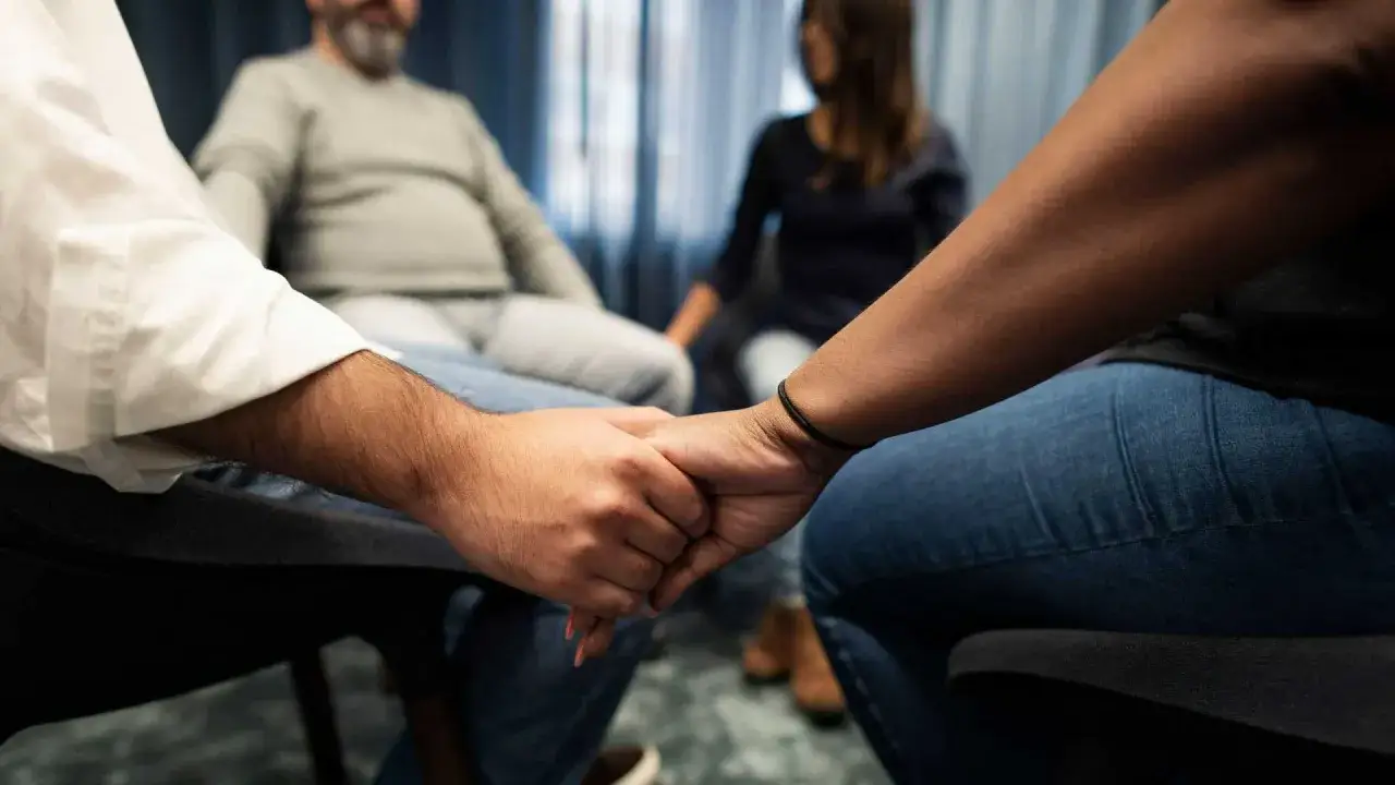 Persone sedute in cerchio si tengono per mano, cercando supporto. Forse per superare il momento in cui mio marito offende la mia famiglia.