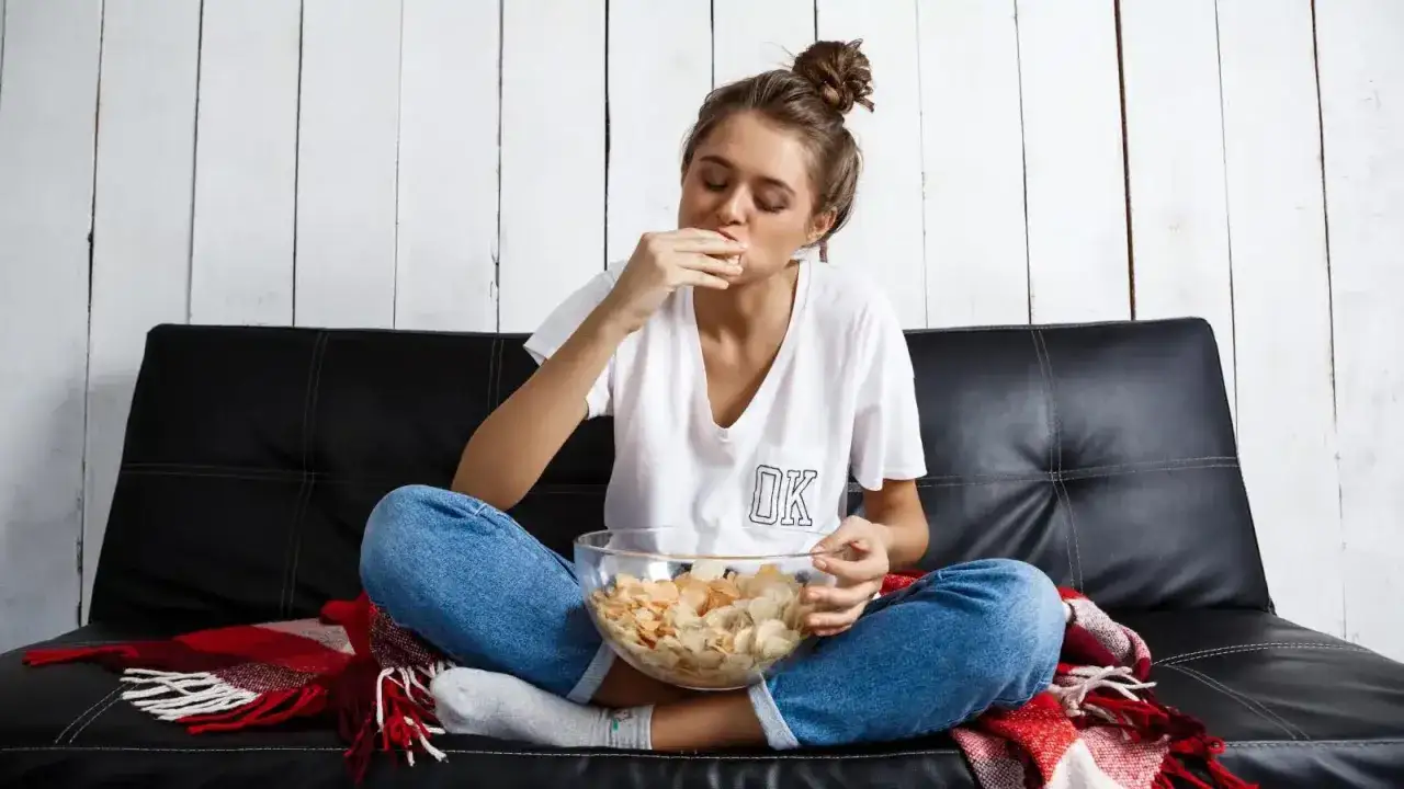Ragazza con ciotola di patatine, sembra avere paura di mangiare, seduta sul divano con una coperta rossa.