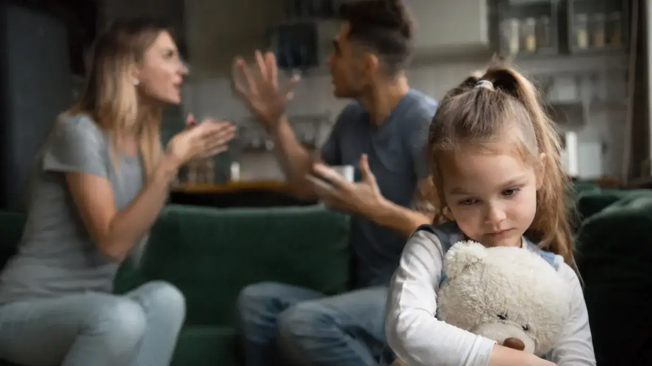 Bambina triste abbraccia orsacchiotto, mentre i genitori litigano sullo sfondo. Crescere in una famiglia dove si litiga sempre è difficile.