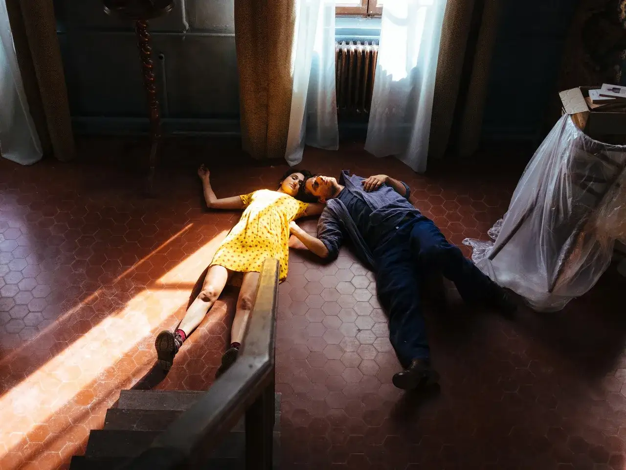 Coppia sdraiata sul pavimento, lei in giallo, lui in blu. La luce filtra dalla finestra, ma lei non accetta la morte di mio marito.