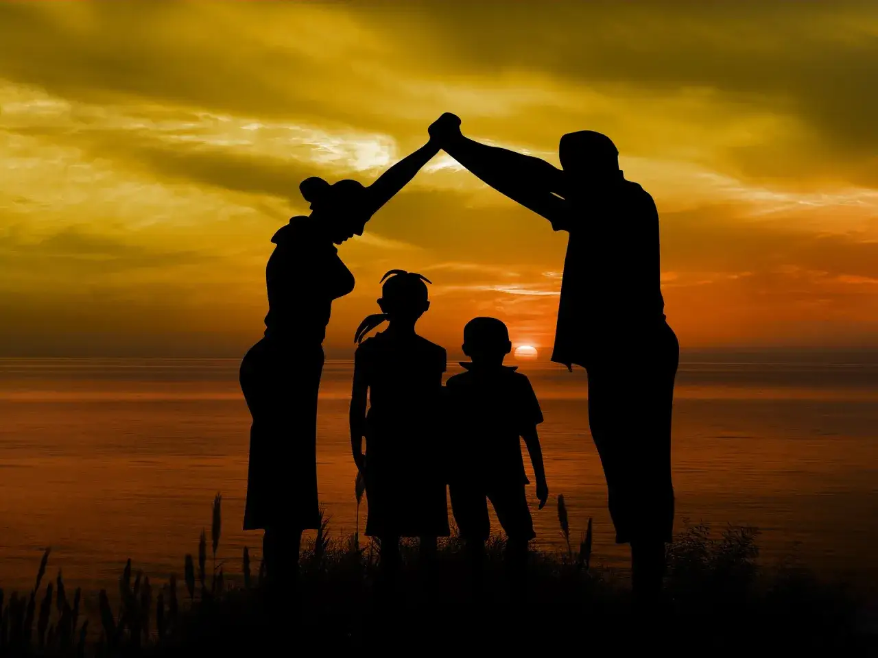 Silhouette di famiglia sotto un tetto di mani, al tramonto sul mare. Riflessioni sui figli che crescono, un abbraccio protettivo che guarda al futuro.