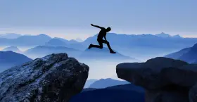 Silhouette di una persona che salta tra due rocce, con montagne blu sullo sfondo. Un viaggio per perdersi per ritrovarsi, un salto nel vuoto per una coppia.
