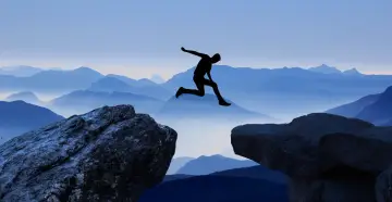 Silhouette di una persona che salta tra due rocce, con montagne blu sullo sfondo. Un viaggio per perdersi per ritrovarsi, un salto nel vuoto per una coppia.
